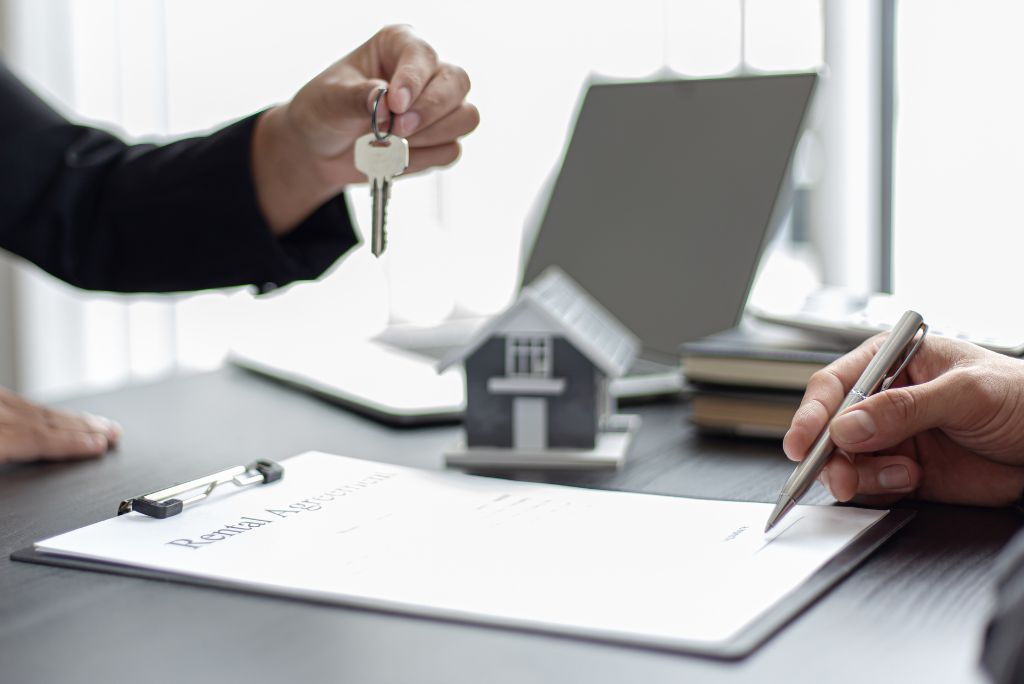 Person handing over a set of keys while another signs a rental agreement document at a desk with a model house and laptop nearby.